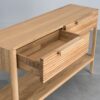 Tento odkládací stolek z masivního dubu kombinuje funkčnost s elegantním designem. Má dvě praktické zásuvky s charakteristickým drážkovaným čelem a minimalistickými úchyty, které dodávají moderní vzhled.