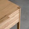 Tento odkládací stolek z masivního dubu kombinuje funkčnost s elegantním designem. Má dvě praktické zásuvky s charakteristickým drážkovaným čelem a minimalistickými úchyty, které dodávají moderní vzhled.